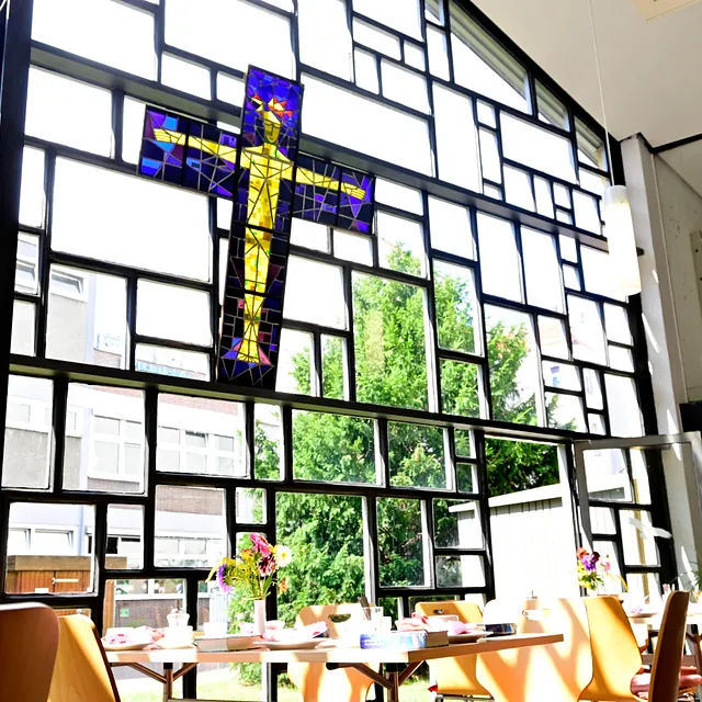 Gemeindesaal mit großem Fenster und Kreuzkunstwerk.